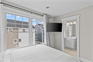 A bedroom with a large window, a flat-screen TV, a bathroom with a sink and mirror, and a patterned bedspread.