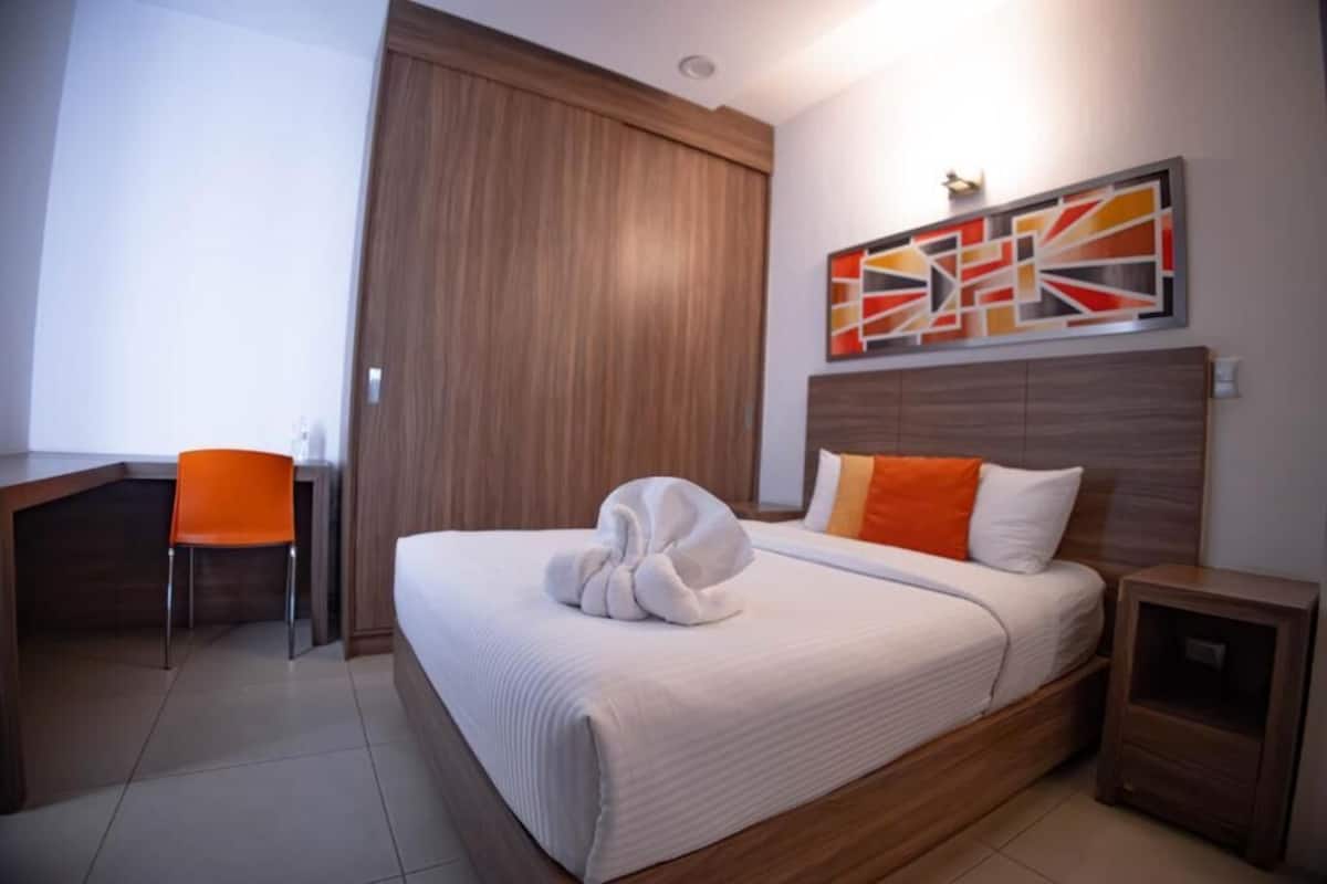 Una habitación de hotel con una cama, un escritorio, un sillón naranja, cabecera de madera y una obra de arte en la pared.