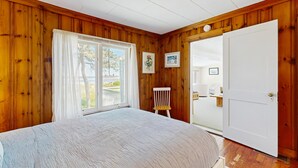 A bedroom with wooden walls, a large bed, a chair, a door, and a window with curtains. - Ocean and Sunset Views: semi private sandy beach, close to dining & Route 1 (Biddeford)