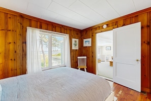 Ein Schlafzimmer mit Holzwänden, einem großen Bett, einem Sessel, einer Tür und einem Fenster mit Vorhängen.