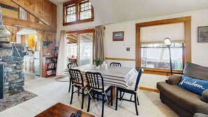 A cozy living room with a stone fireplace, a dining table with chairs, a sofa with a cushion, and a large window with a view.