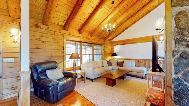 Interior de una cabaña de troncos con techo de madera, un sofá de cuero, un sofá seccional blanco, una mesa de centro de madera y una chimenea de piedra.