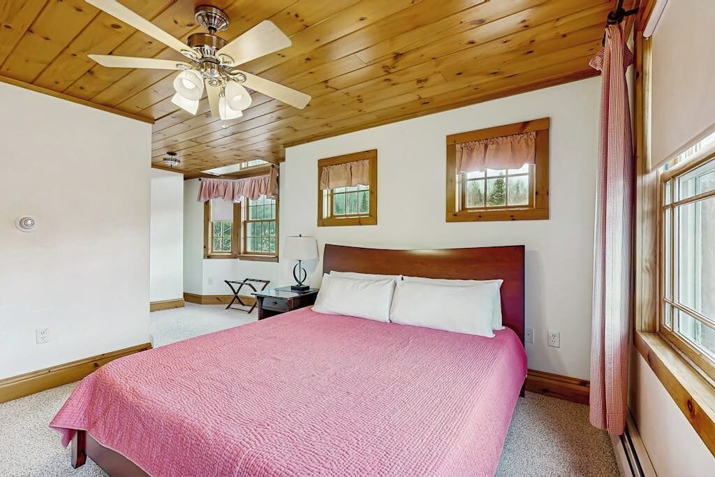Un dormitorio con techo de madera, una cama con manta rosa, una mesita de noche, un escritorio con silla y una ventana con cortinas.