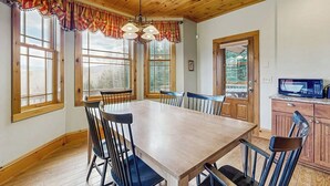 Uma sala de jantar com mesa e cadeiras de madeira, janelas amplas e uma área de cozinha com micro-ondas.