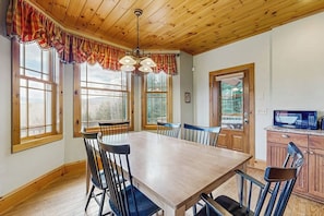 A dining area with a wooden table and chairs, large windows, and a kitchen area with a microwave.