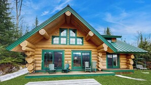 Une cabane en rondins avec un toit végétalisé, de grandes fenêtres et une terrasse avec des chaises.