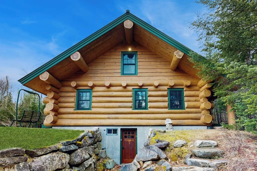 A log cabin with a green roof, a central chimney, and a stone foundation.