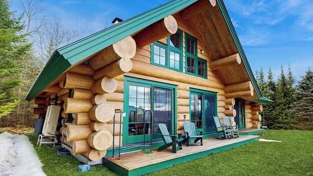 Une cabane en rondins dotée d’une véranda, de grandes fenêtres et d’une pile de bois à l’extérieur.