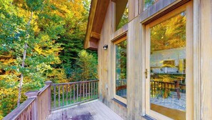 Une terrasse en bois avec une balustrade, de grandes fenêtres et une vue sur une zone boisée.