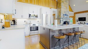 Une cuisine moderne avec des meubles blancs, un îlot central doté de tabourets, une cheminée en pierre et un téléviseur fixé au mur.