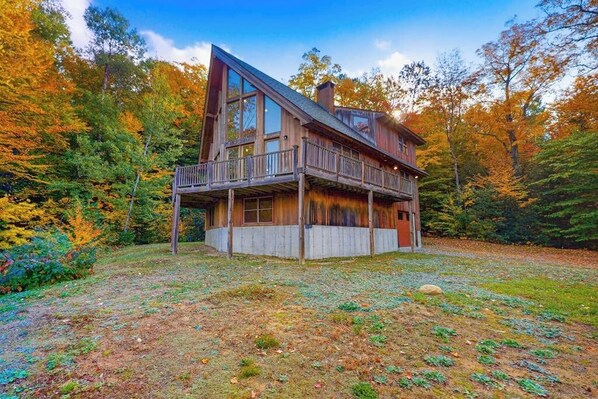 A two-story wooden cabin with a balcony, surrounded by autumn foliage.