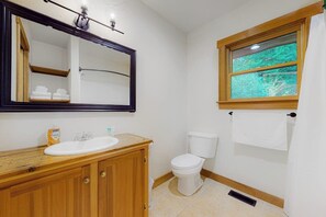 A bathroom with a wooden vanity, a white sink, a mirror, a toilet, and a window with a view of greenery.