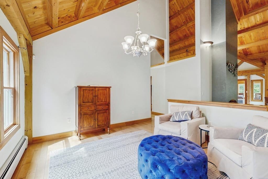 A spacious room with wooden ceiling beams, a chandelier, a white sofa, a blue ottoman, and a wooden cabinet.