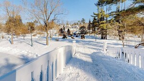 Paisaje nevado con una valla blanca, árboles y un edificio al fondo.