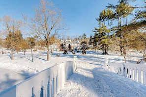 A snow-covered landscape with a white fence, trees, and a building in the background.
