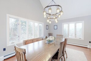 Ein Esszimmer mit einem großen Holztisch, sechs Stühlen, einer Chandelierleuchtre und großen Fenstern mit Blick auf Bäume.