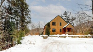 Una casa de madera de dos pisos con cimientos de piedra, rodeada de terreno y árboles cubiertos de nieve.
