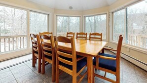 Un comedor con mesa y sillas de madera, ventanas amplias y vista a los árboles.
