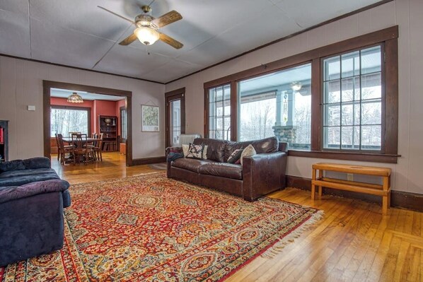 A spacious living room with a large wooden floor, a brown leather sofa, a dark blue armchair, a wooden bench, and a patterned rug. - Cozy home with  covered porch, & sun room - walk to shops/dining/bars (Bethlehem)