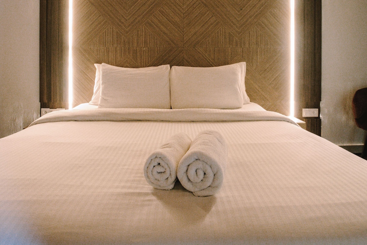 A hotel room with a large bed, a wooden headboard, and two rolled towels on the bed.