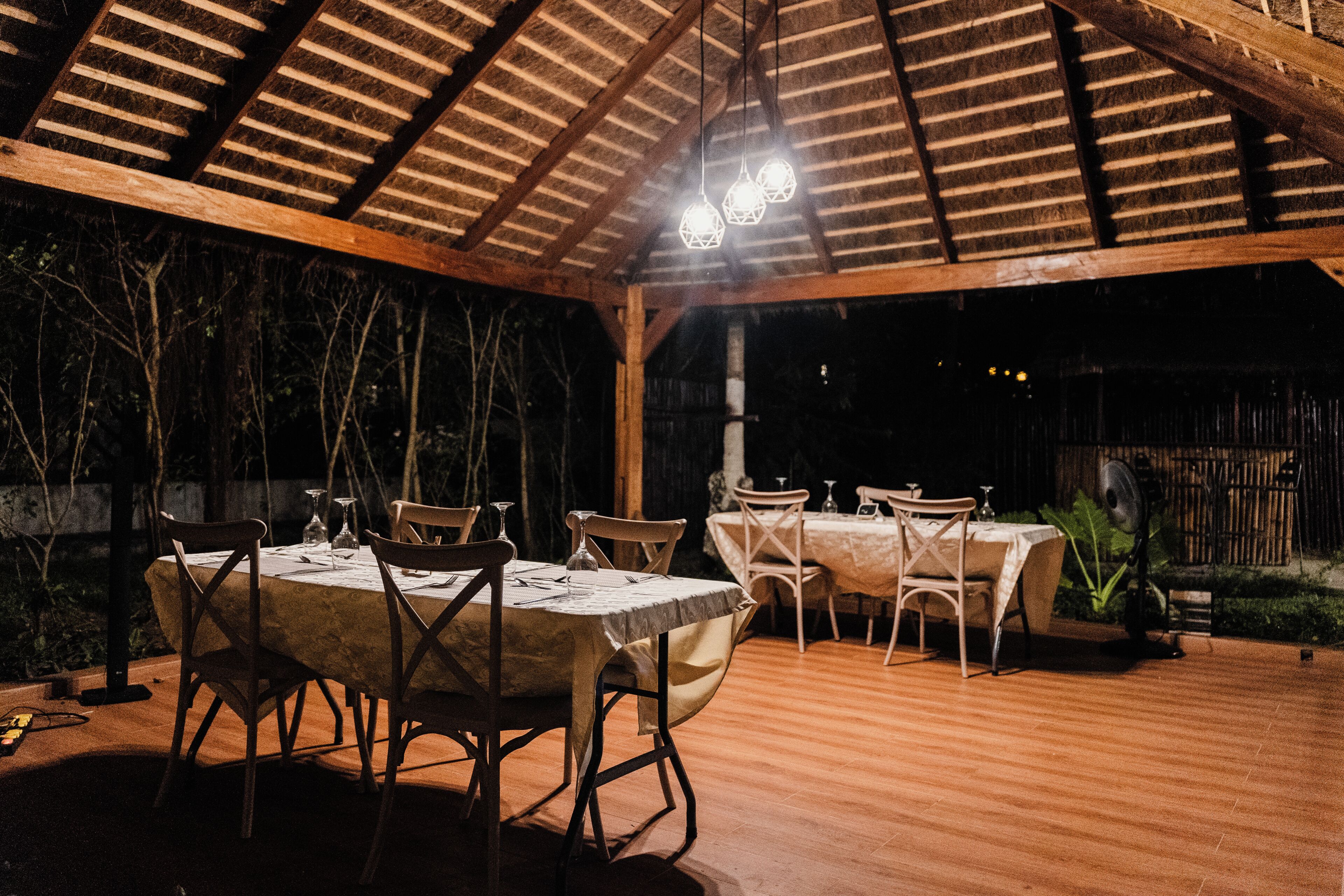 Un patio al aire libre con mesas y sillas de madera, techo de paja y luces colgantes.