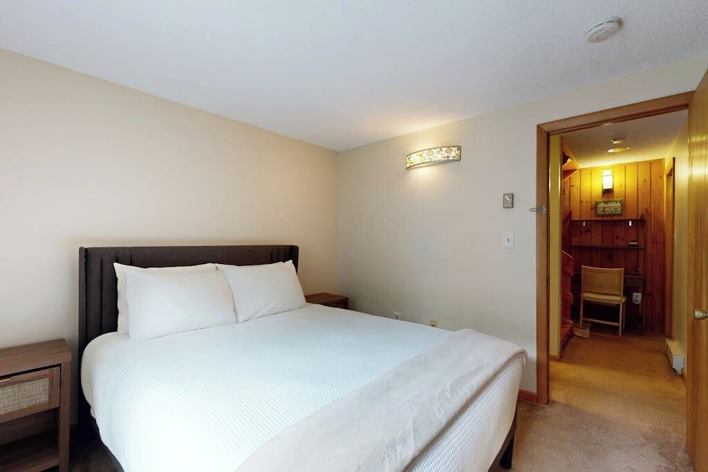 A bedroom with a bed, bedside tables, a wall-mounted light, and a door leading to a wooden room.