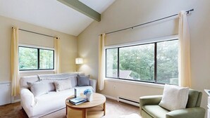 Ein Wohnzimmer mit einer Couch, einem Sessel, einem Couchtisch und großen Fenstern mit Vorhängen.