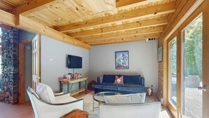 Una sala de estar acogedora con pared de piedra, techo de madera, televisor de pantalla plana, un sofá azul y un sillón blanco.