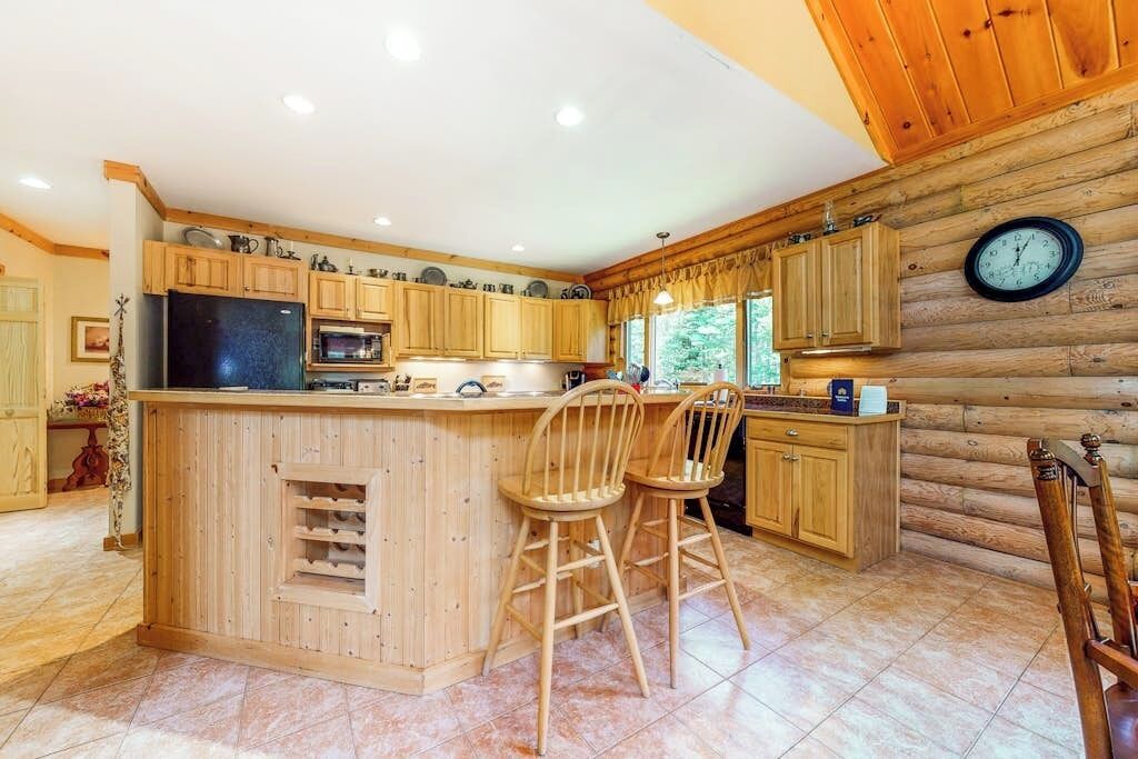 Een keuken in blokhutstijl met houten kasten, een centraal eiland met barstoelen, een koelkast, magnetron en een klok aan de muur.