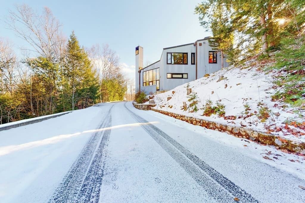 A snow-covered road leading to a modern house with large windows and a stone retaining wall.