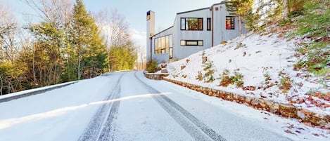 Une route enneigée menant à une maison moderne dotée de grandes fenêtres et d’un mur de soutènement en pierre.