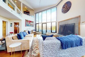 Ein geräumiges Wohnzimmer mit hoher Decke, einem großen Fenster, einer Sofagarnitur, einem Bett und einem Fernseher.