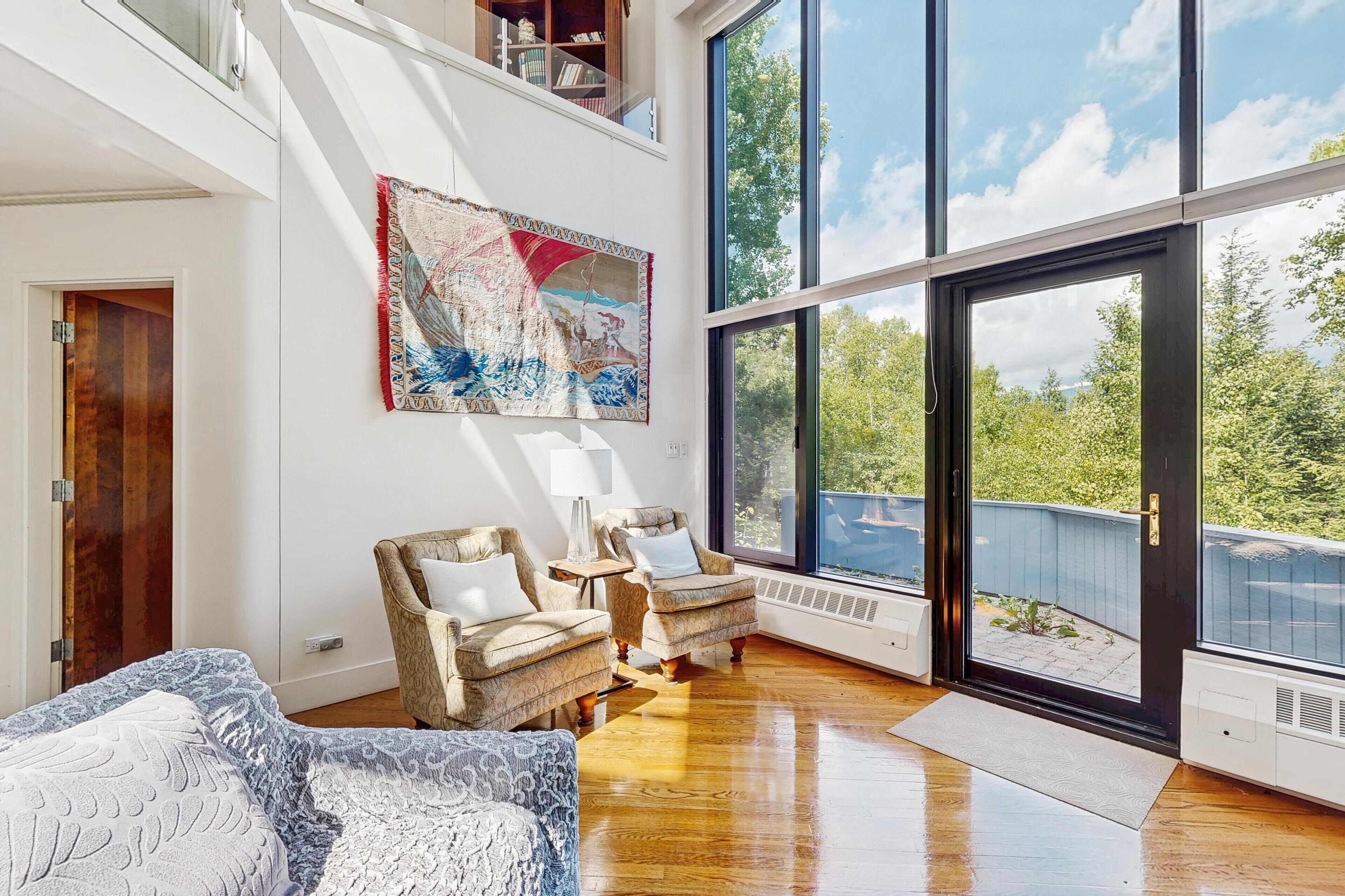 A spacious living room with a high ceiling, large windows, and a view of greenery outside.