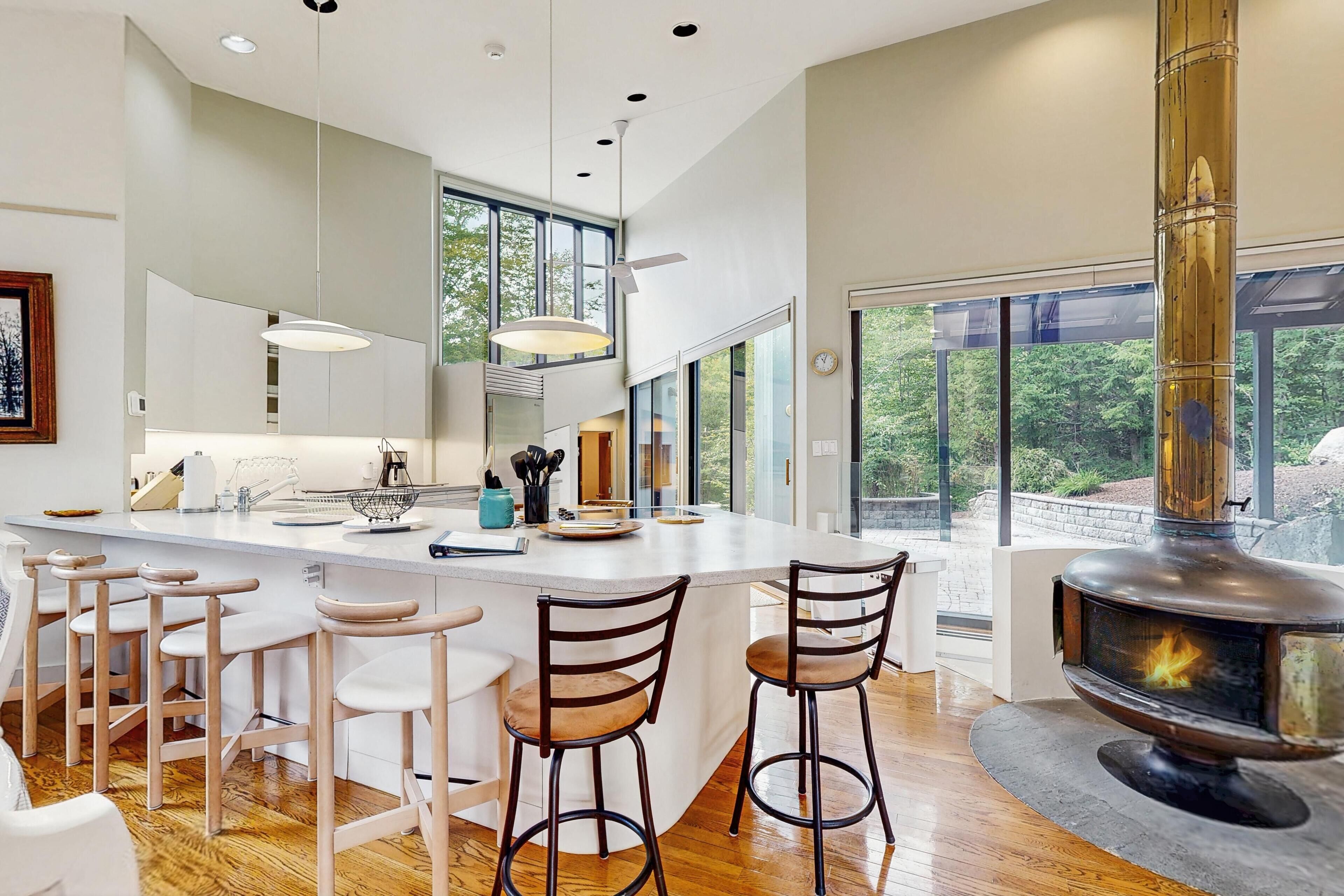 A modern kitchen with a large island, bar stools, a wood-burning stove, and large windows offering a view of the outdoors.