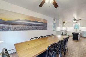 Un espace repas avec une longue table en bois et des chaises, une cuisine avec des meubles blancs et un grand tableau représentant un paysage accroché au mur.