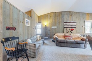 Ein gemütlicher Raum mit einem karierten Sofa, einem gepolsterten Sessel und einem grauen Sofa. Eine Holzwand mit dem Schriftzug 'Black Bear Lodge' ist sichtbar.