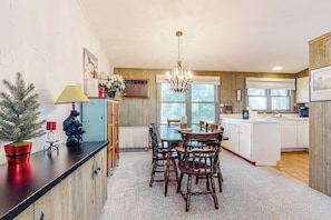 Ein Essbereich mit Holztisch und -stühlen, einer Chandelüster und einer Küche mit weißen Schränken und einem Fenster.