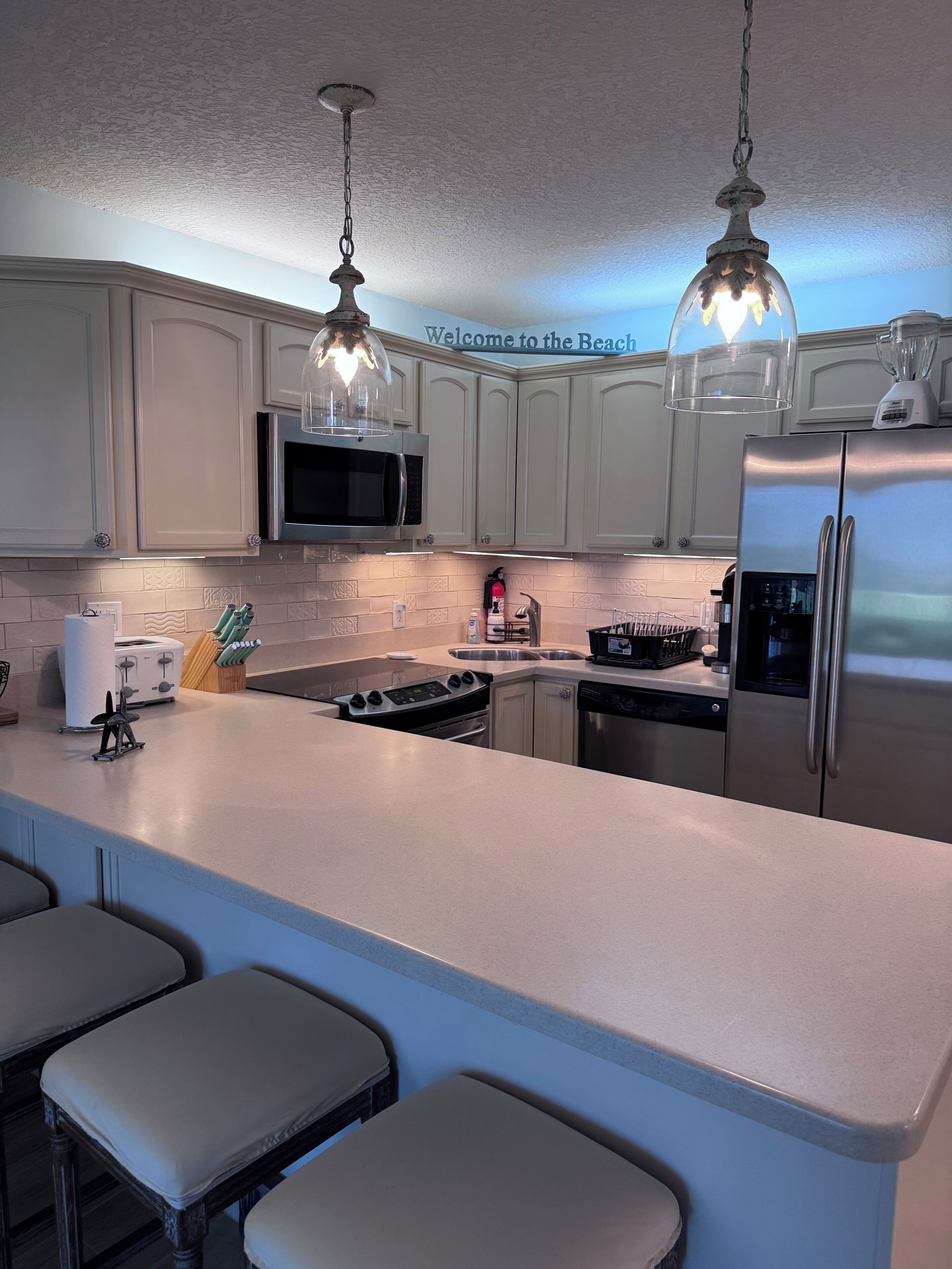 Una cocina moderna con gabinetes blancos, electrodomésticos de acero inoxidable, una isla central con taburetes altos y luminarias colgantes.