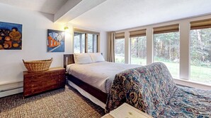 Ein Schlafzimmer mit einem Bett, einer Kommode, einem gemusterten Sofa und großen Fenstern mit Blick auf Bäume.