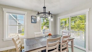 Uma área de refeições com mesa de madeira, cadeiras brancas, lustre e janelas amplas com vista para o verde.