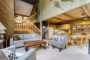 Ein großzügiger Wohnbereich mit einer Couch, einem Couchtisch, einem gemusterten Hocker und einem Holzes Esstisch mit Stühlen. Eine Treppe führt zu einer oberen Ebene.