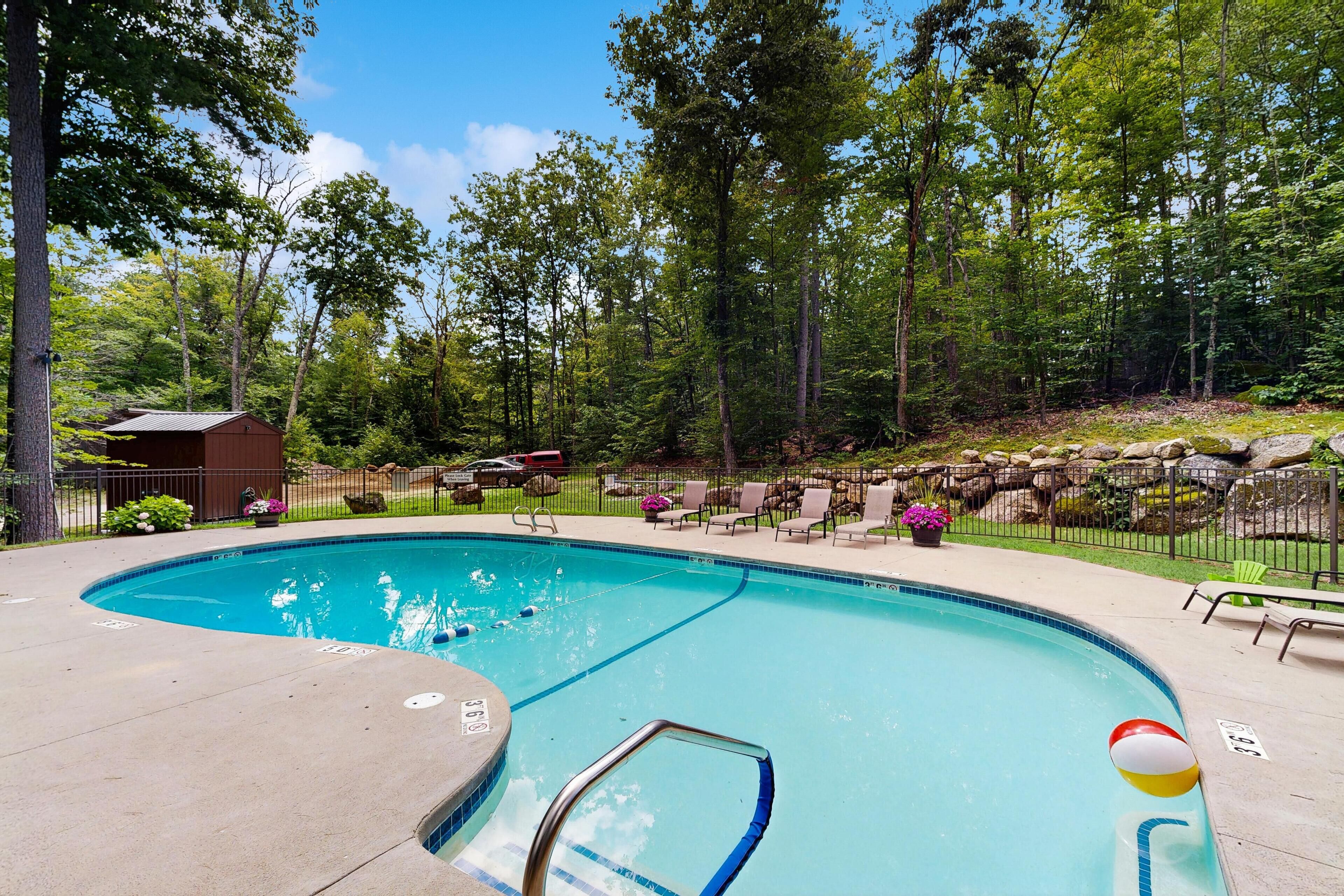 A swimming pool with a curved design, surrounded by a fence, trees, and a small building.
