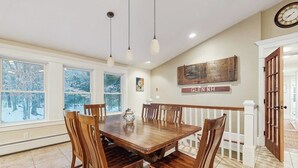 A dining area with a wooden table and chairs, large windows with a view of trees, a clock on the wall, and a sign that reads 'GLEN NH'. - Serene Dog Friendly Home:  near skiing, hiking, Storyland, Shopping & dining (Glen)