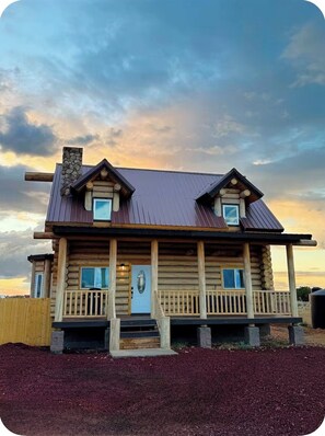 A two-story log cabin with a porch, a stone chimney, and a view of the sky.