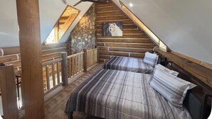 Une chambre située dans un espace de type « loft », avec un plafond en pente, des poutres en bois, un mur en pierre, un lit avec une literie à rayures, un tableau représentant un chien et un ventilateur de plafond.