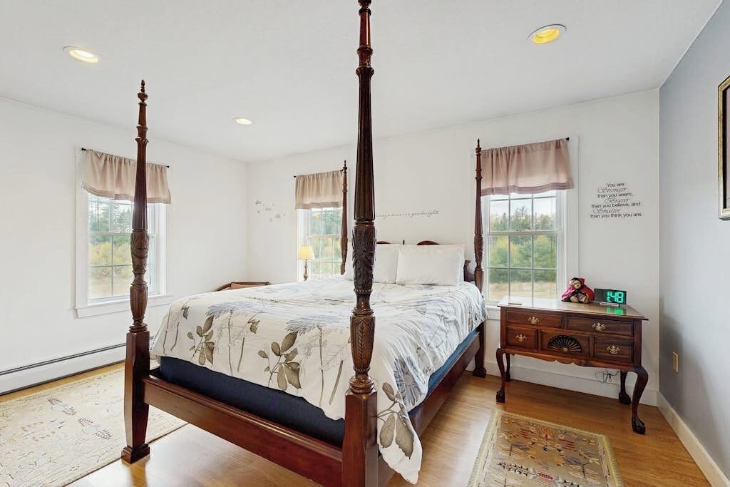 A four-poster bed with a floral bedspread, a wooden nightstand with a clock, and a view of the outdoors through a window.