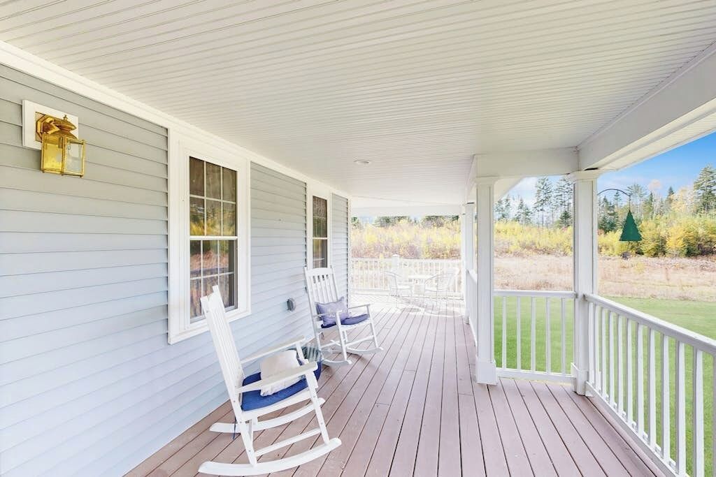 A covered porch with white railings, rocking chairs, and a view of a grassy landscape.