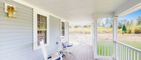 A covered porch with white railings, rocking chairs, and a view of a grassy landscape.