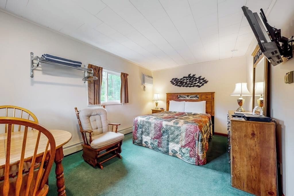 A bedroom with a bed, a chair, a table, a dresser, a mirror, and a wall-mounted TV.