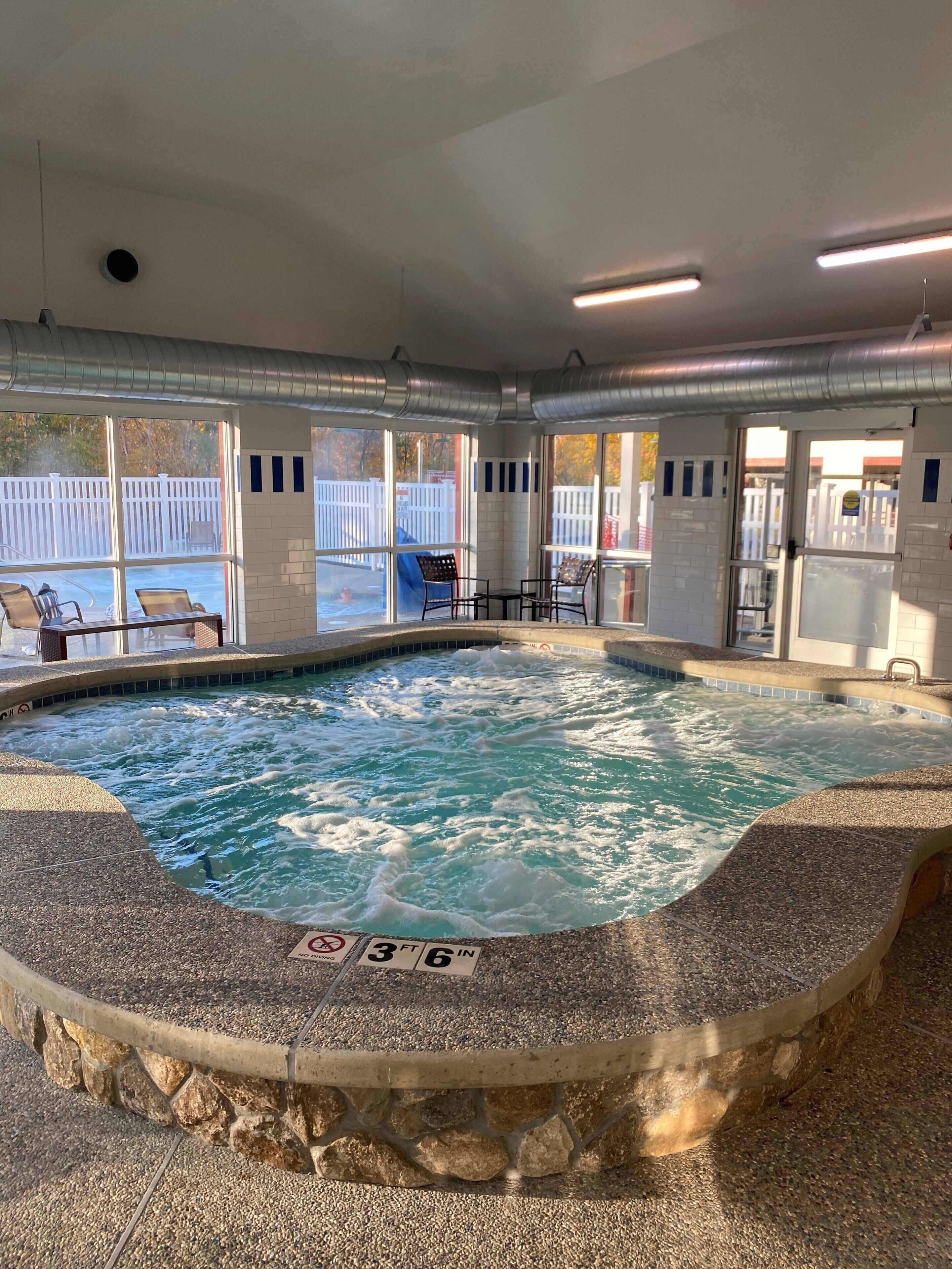 A hot tub with a stone surround, indoor seating area with chairs, and a view of a white fence outside.